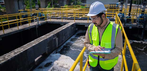 man in yellow vest and white hard hat looks over the edge of a wastewater treatment pond