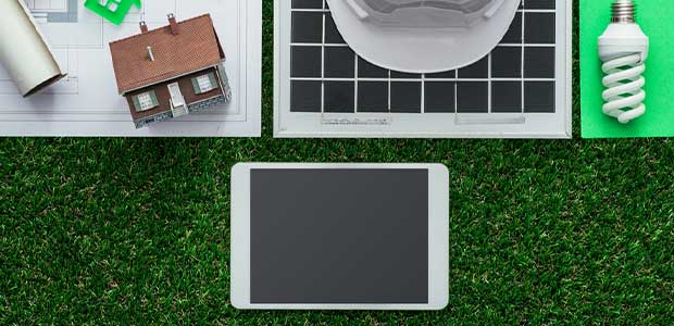 a digital tablet sits on a grassy backdrop with a hard hat, solar panel, model of a home, lightbulb and blueprint above